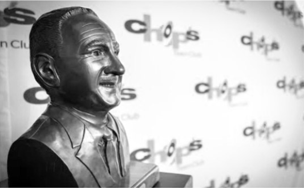 Bronze bust of a man in a suit, smiling and looking right. Background has repeating words "Chops" and "Vision Club.